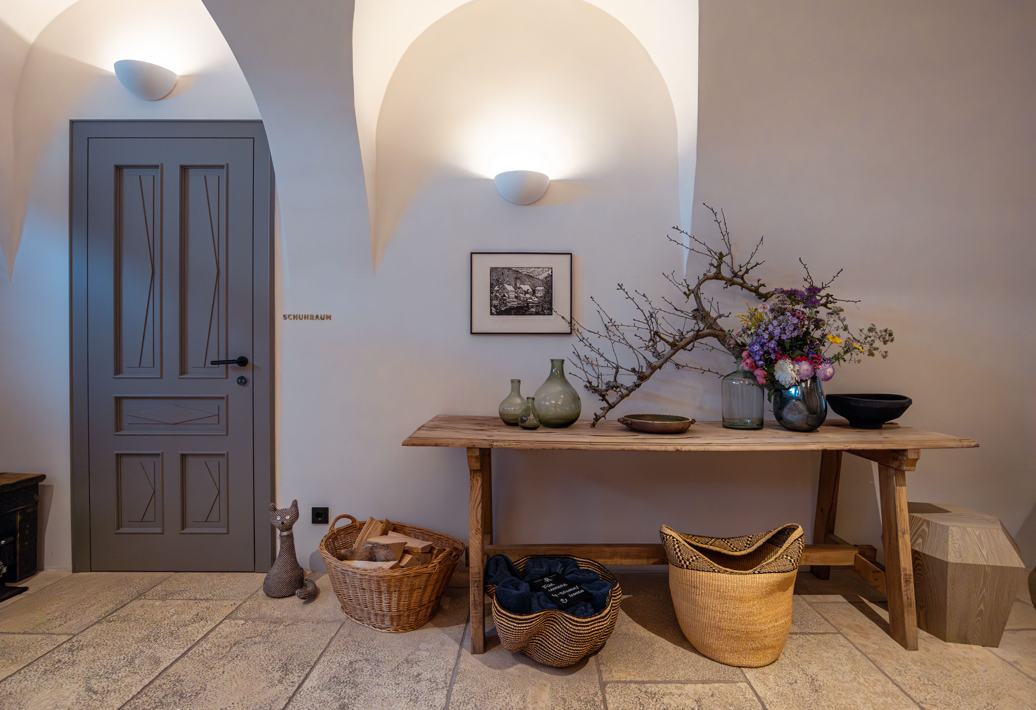 Innenraum mit Holztisch, Vasen, Blumen und Körben. Tür mit Aufschrift 'Schuhraum'.