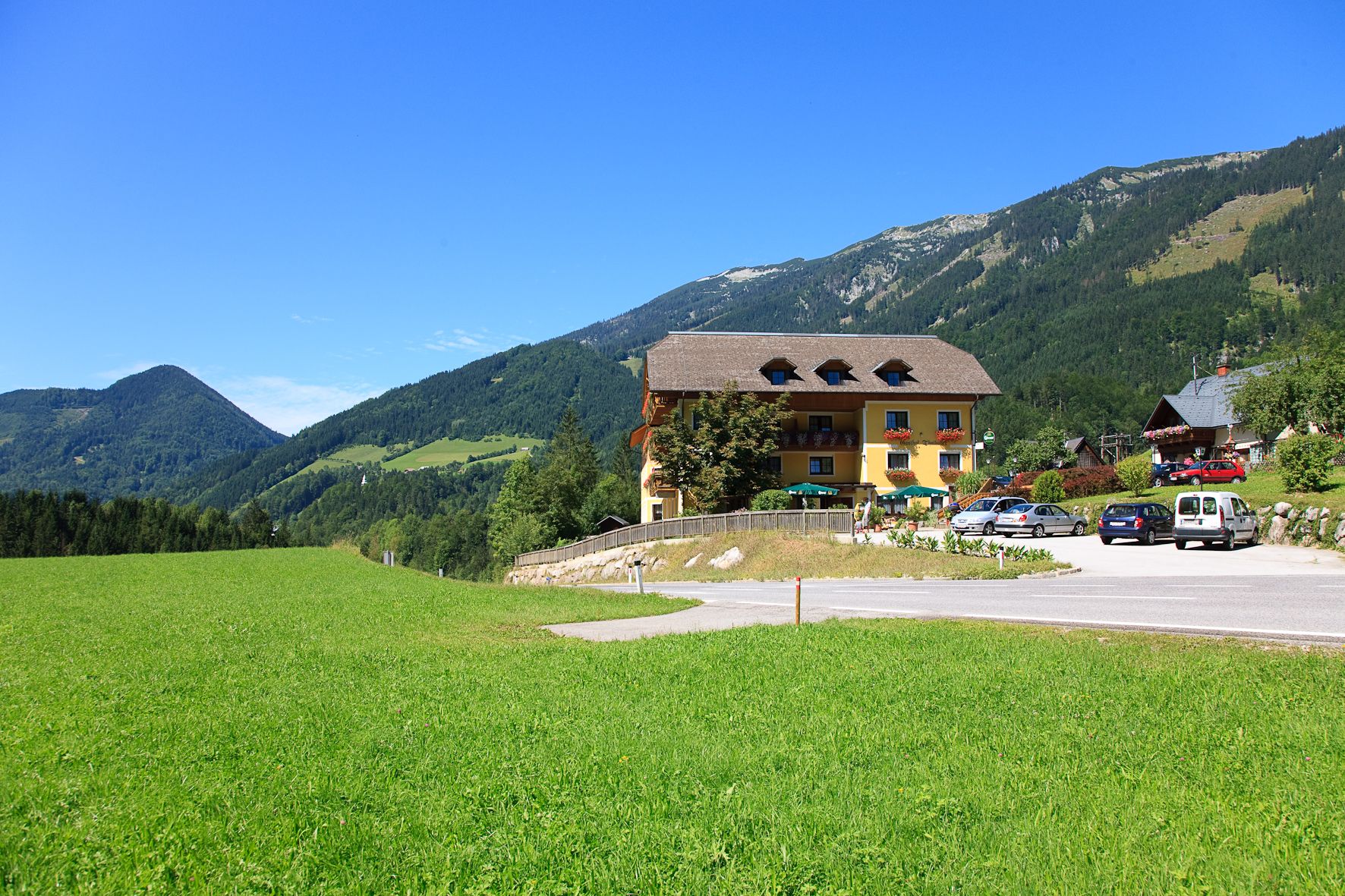 Ein gelbes Gebäude mit Balkon und Blumen, umgeben von grünen Wiesen und Bergen unter blauem Himmel.