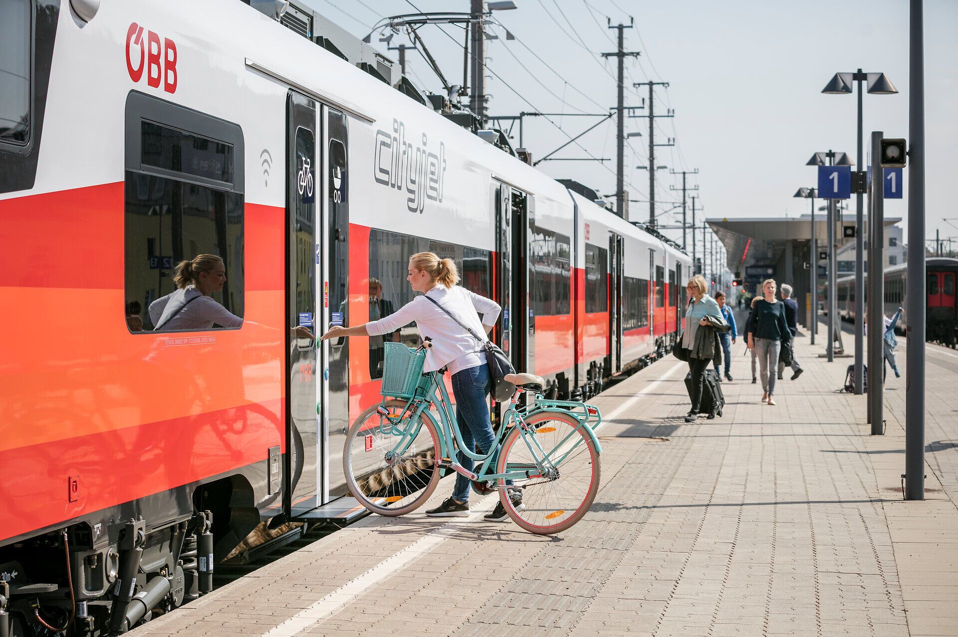 Am Bahnhof herrscht reges Treiben, während Reisende mit ihren Fahrrädern auf den Zug warten. Die Sonne strahlt am klaren Himmel und verspricht einen perfekten Tag für Abenteuer in der Natur.