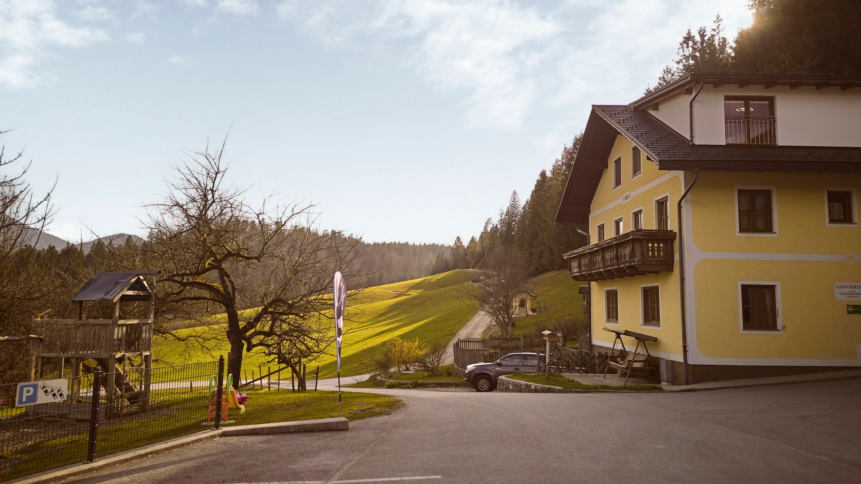Der Familienbauernhof Glockriegl- ein gelbes Bauernhaus mit Balkon steht in einer grünen, hügeligen Landschaft.