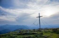 Ein majestätisches Gipfelkreuz thront über den sanften Hügeln, umgeben von einer atemberaubenden Berglandschaft. Die Wolken ziehen sanft über den Himmel und verleihen der Szenerie eine friedliche Atmosphäre, während die frische Bergluft zum Wandern einlädt.