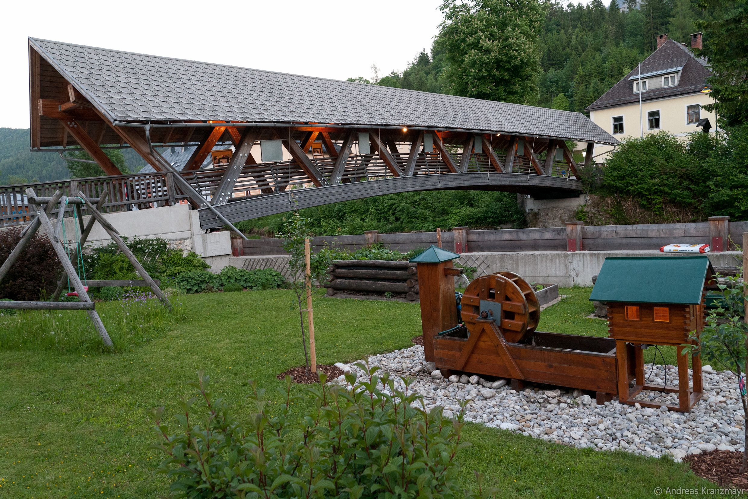 Überdachte Holzbrücke über einen Fluss mit einem kleinen Wasserrad im Vordergrund.
