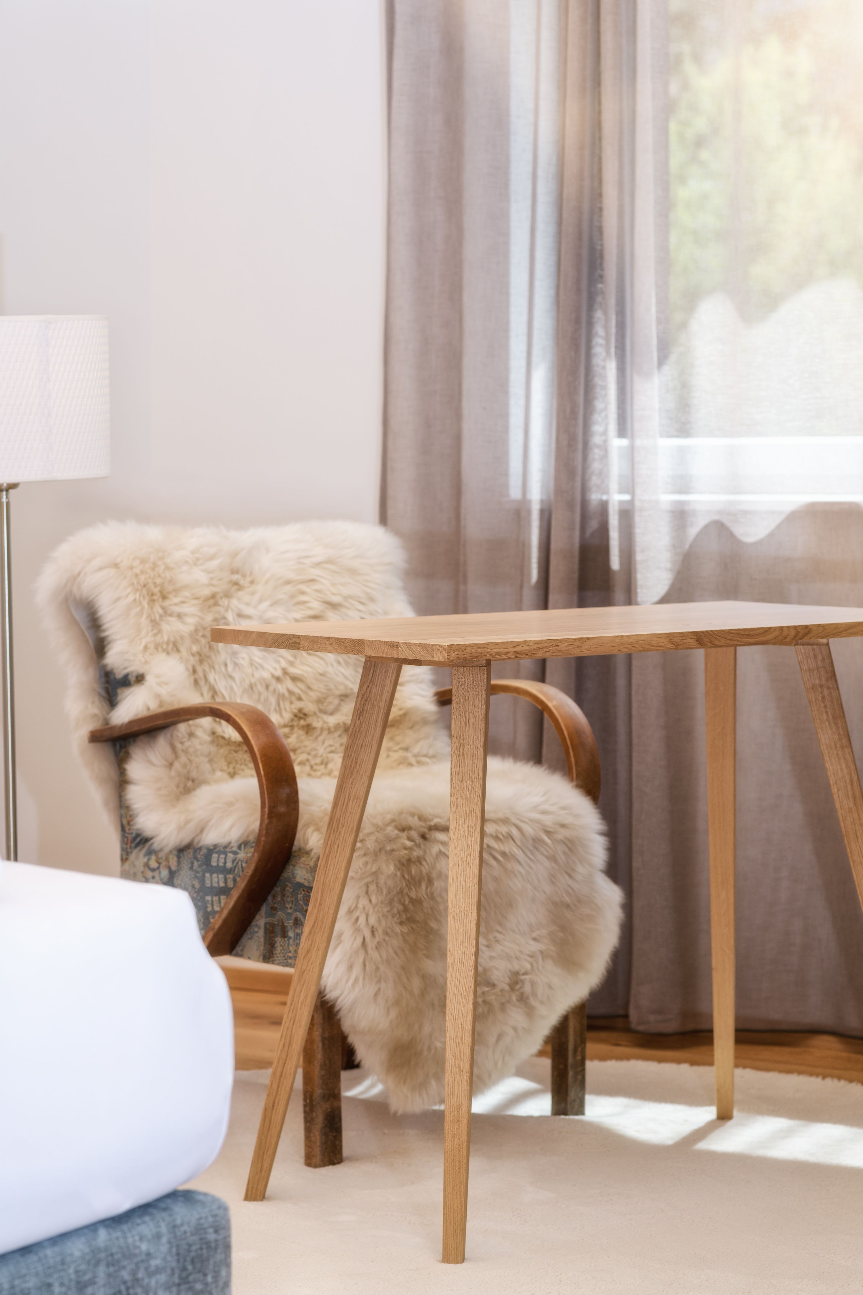 Ein gemütlicher Raum mit einem Holztisch, einem Stuhl mit Fellüberwurf und einem Fenster mit Vorhängen.