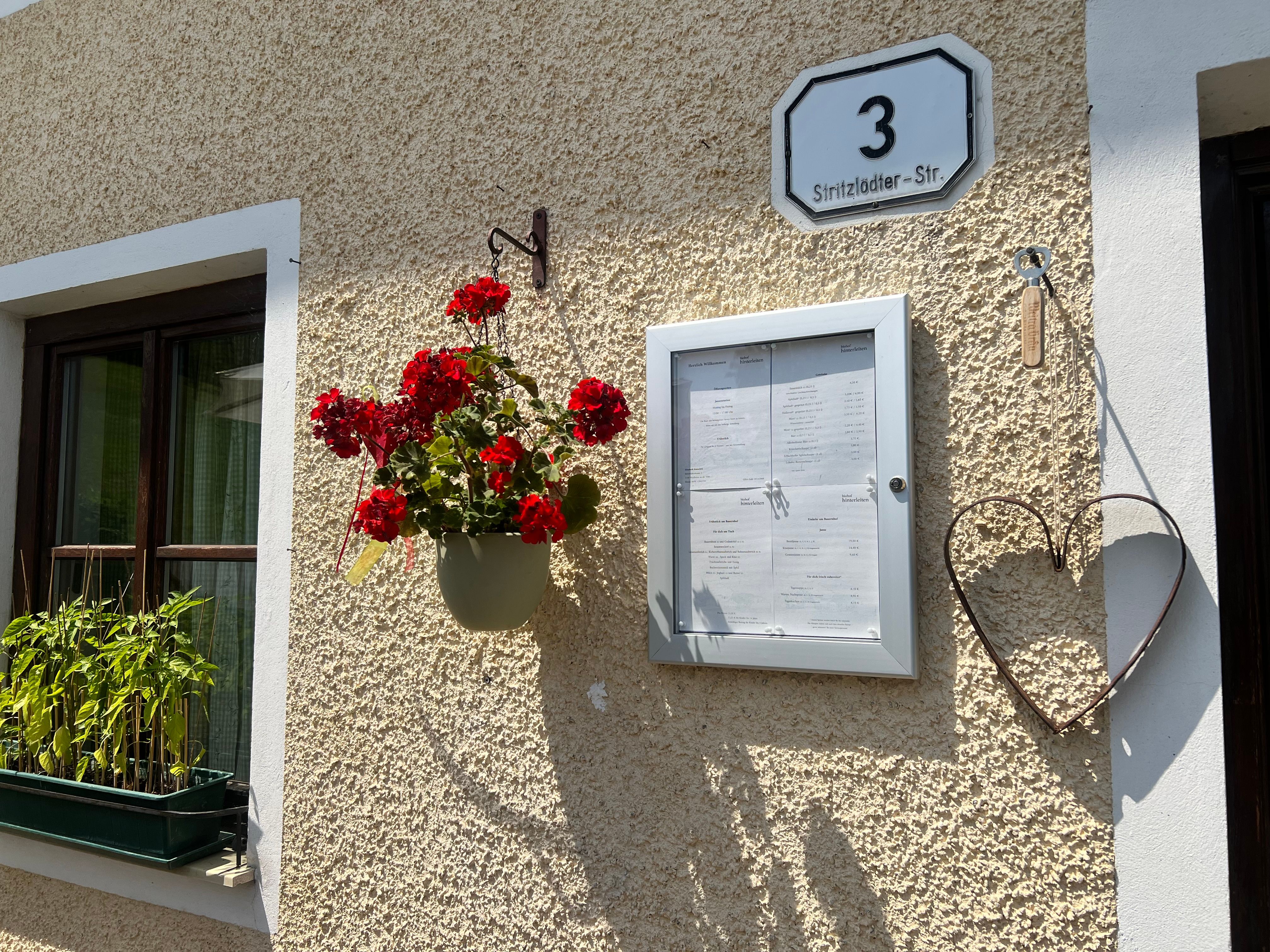 Außenansicht einer Hauswand mit einer Adresse, einem Blumenarrangement und einem Herz aus Metall.