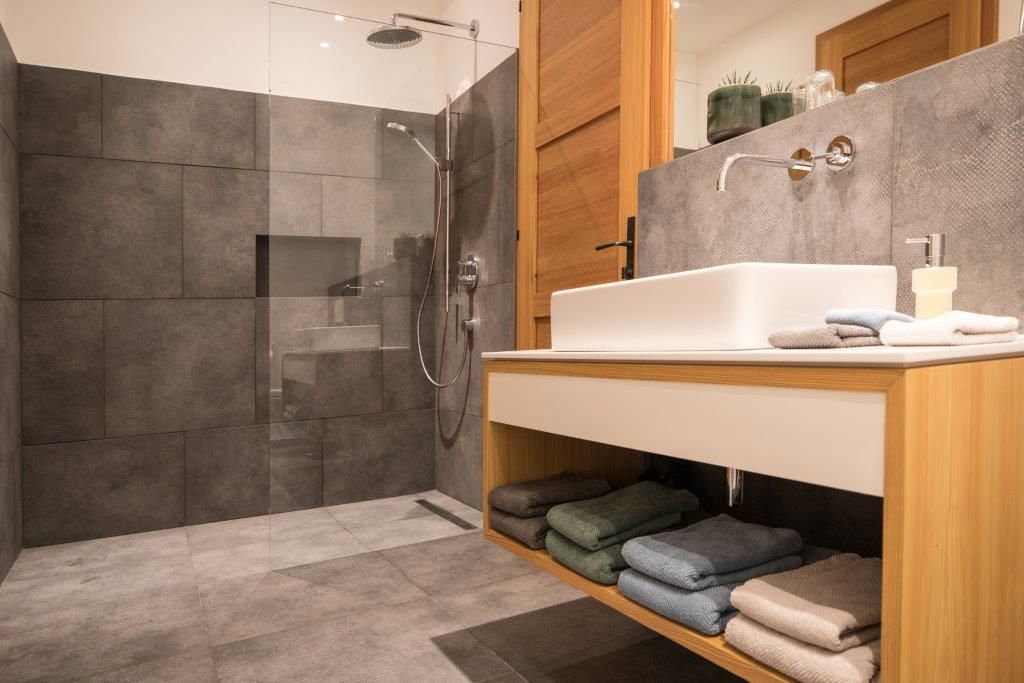 Modernes Badezimmer mit grauen Fliesen, einer Glasdusche und einem Holzwaschtisch mit Handtüchern.