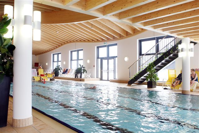 Innenansicht eines Schwimmbads mit Holzdecke und großen Fenstern.