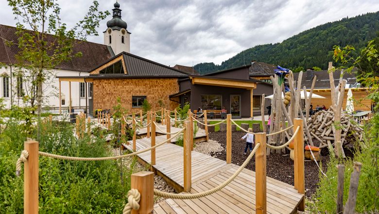 Ein modernes Gebäude mit Holzverkleidung und Spielplatz, umgeben von grüner Landschaft und Bergen im Hintergrund.