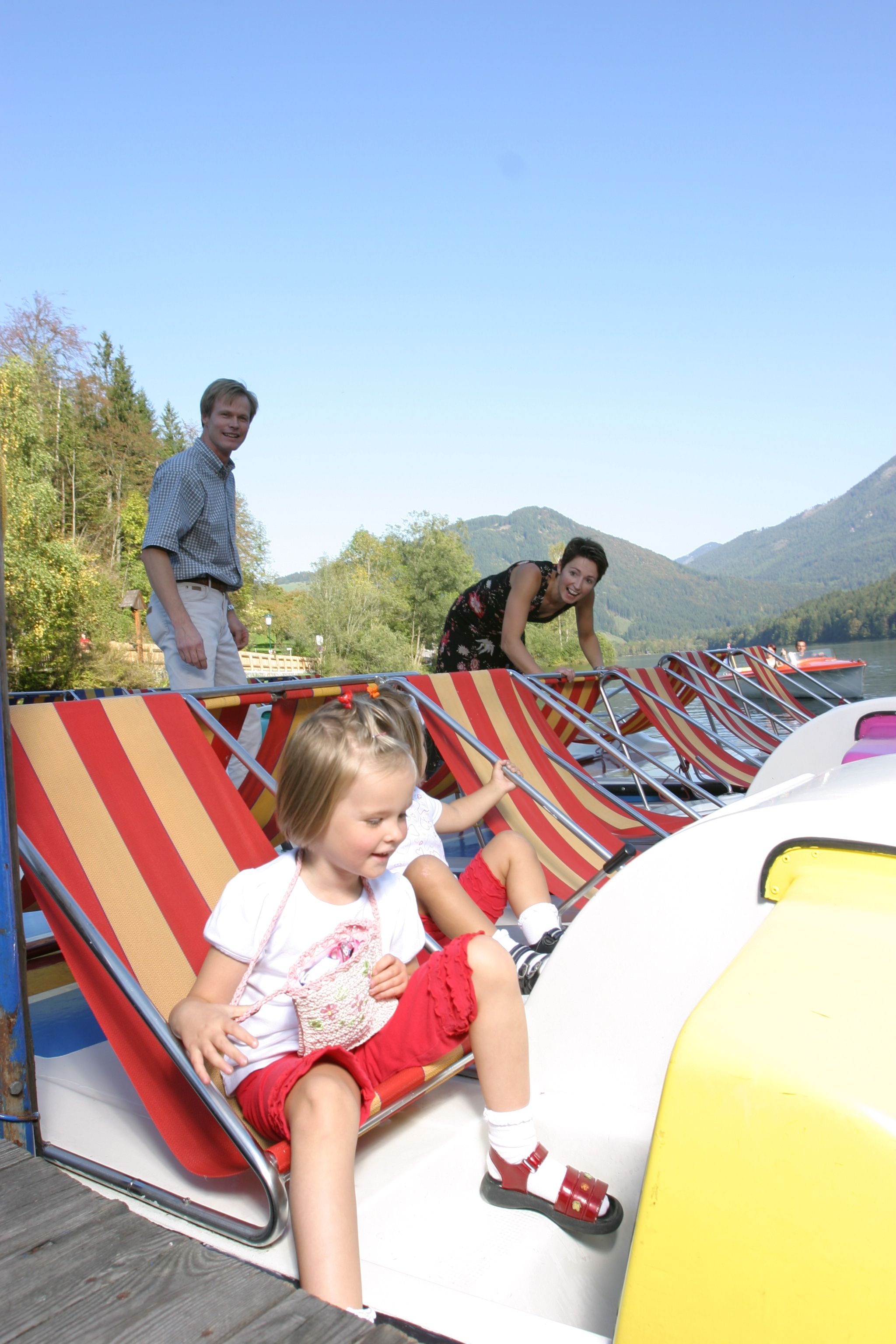 Familie mit Kindern auf Tretbooten an einem See mit Bergen im Hintergrund.