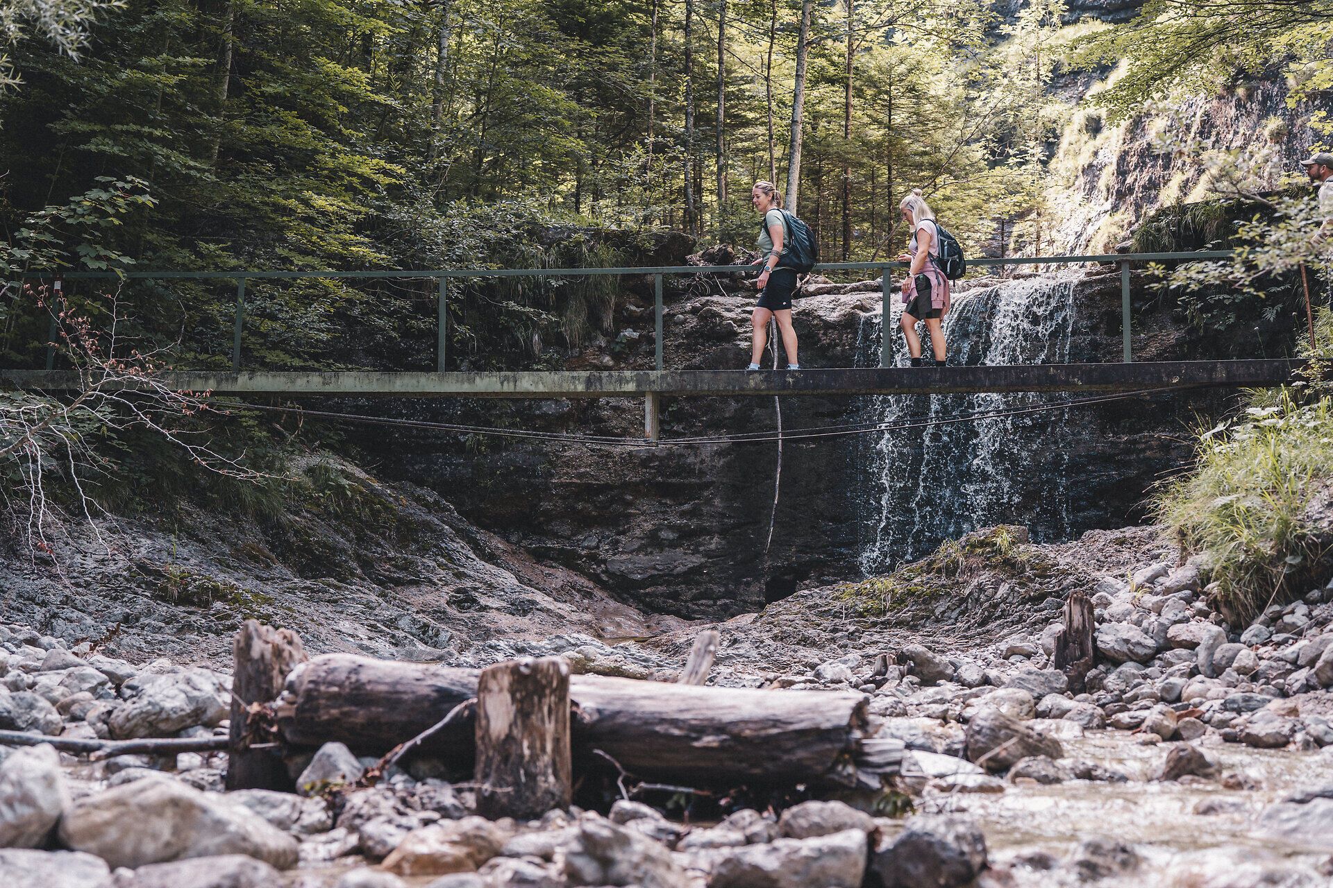 Zwei Wandernde überqueren eine schmale Brücke vor einem kleinen Wasserfall in einer bewaldeten Schlucht, darunter ein steiniger Bachlauf mit Treibholz im Vordergrund.