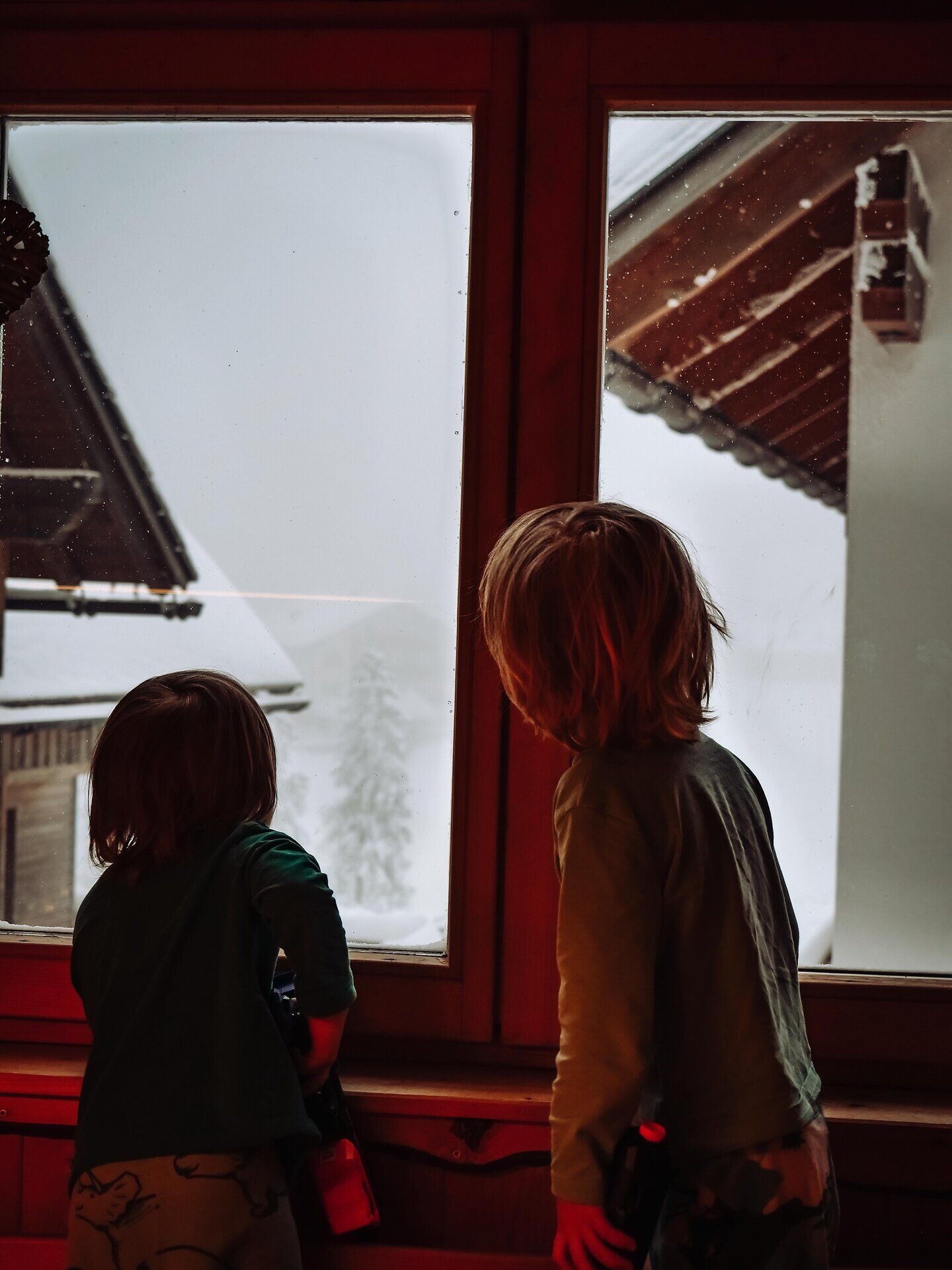 Zwei Kinder stehen fasziniert am Fenster und beobachten den sanften Schneefall, der die Landschaft in eine traumhafte Winterwelt verwandelt. Die gemütliche Atmosphäre des Raumes lädt dazu ein, die kalte Jahreszeit in vollen Zügen zu genießen und die Schönheit der verschneiten Berge zu bewundern.