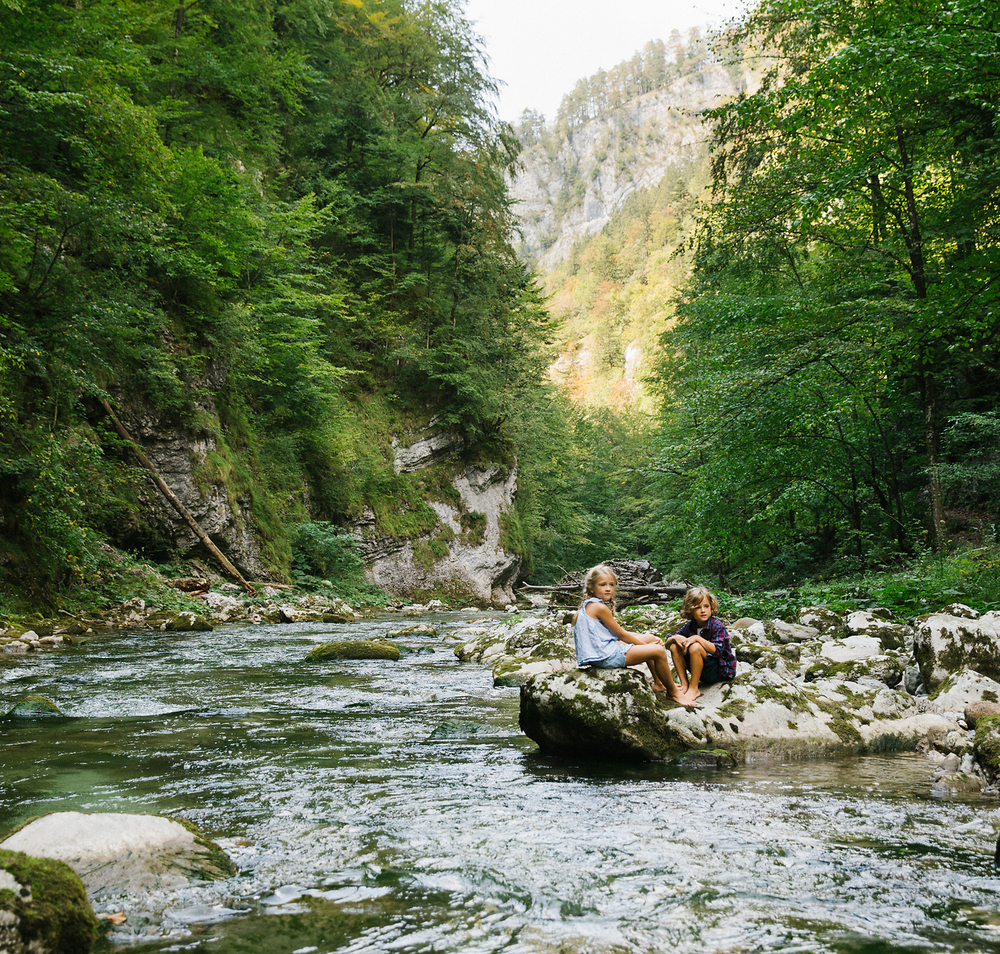 Ein erfrischender Fluss schlängelt sich durch eine grüne Landschaft, umgeben von hohen Bäumen und sanften Hügeln. Familien genießen die Ruhe der Natur, während sie auf den glatten Steinen am Ufer sitzen und die kühle Brise spüren. Hier wird der Sommer in den Bergen zu einem unvergesslichen Erlebnis.
