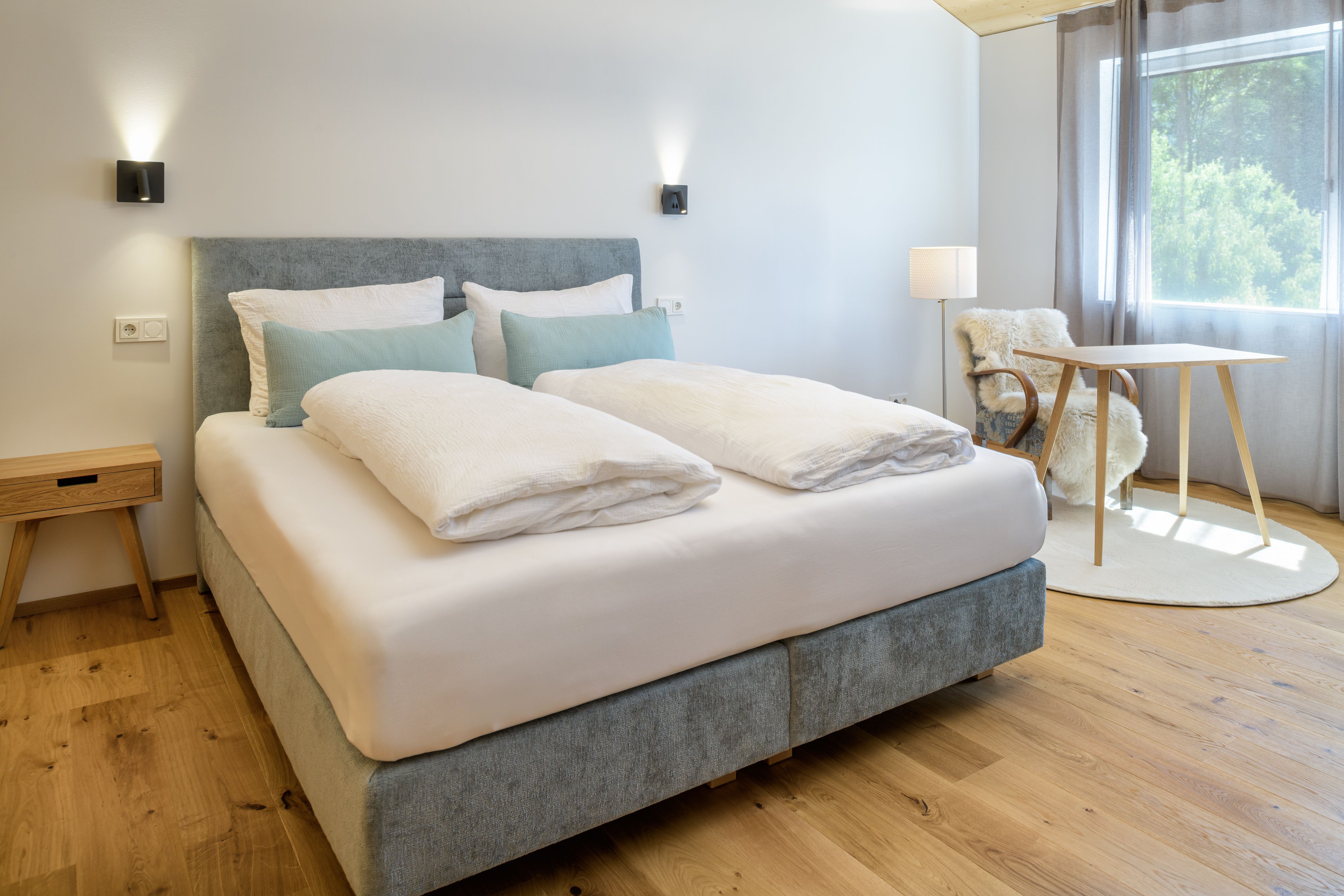 Ein modernes Schlafzimmer mit einem großen Bett, Holzboden, einem kleinen Tisch und einem Stuhl mit Fellüberwurf. Tageslicht strömt durch ein Fenster.