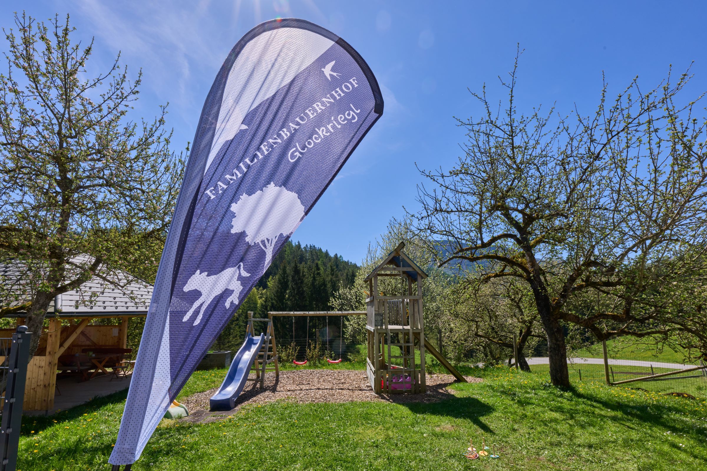 Spielplatz mit Rutsche und Schaukel im Garten eines Bauernhofs, umgeben von Bäumen und Wiesen.