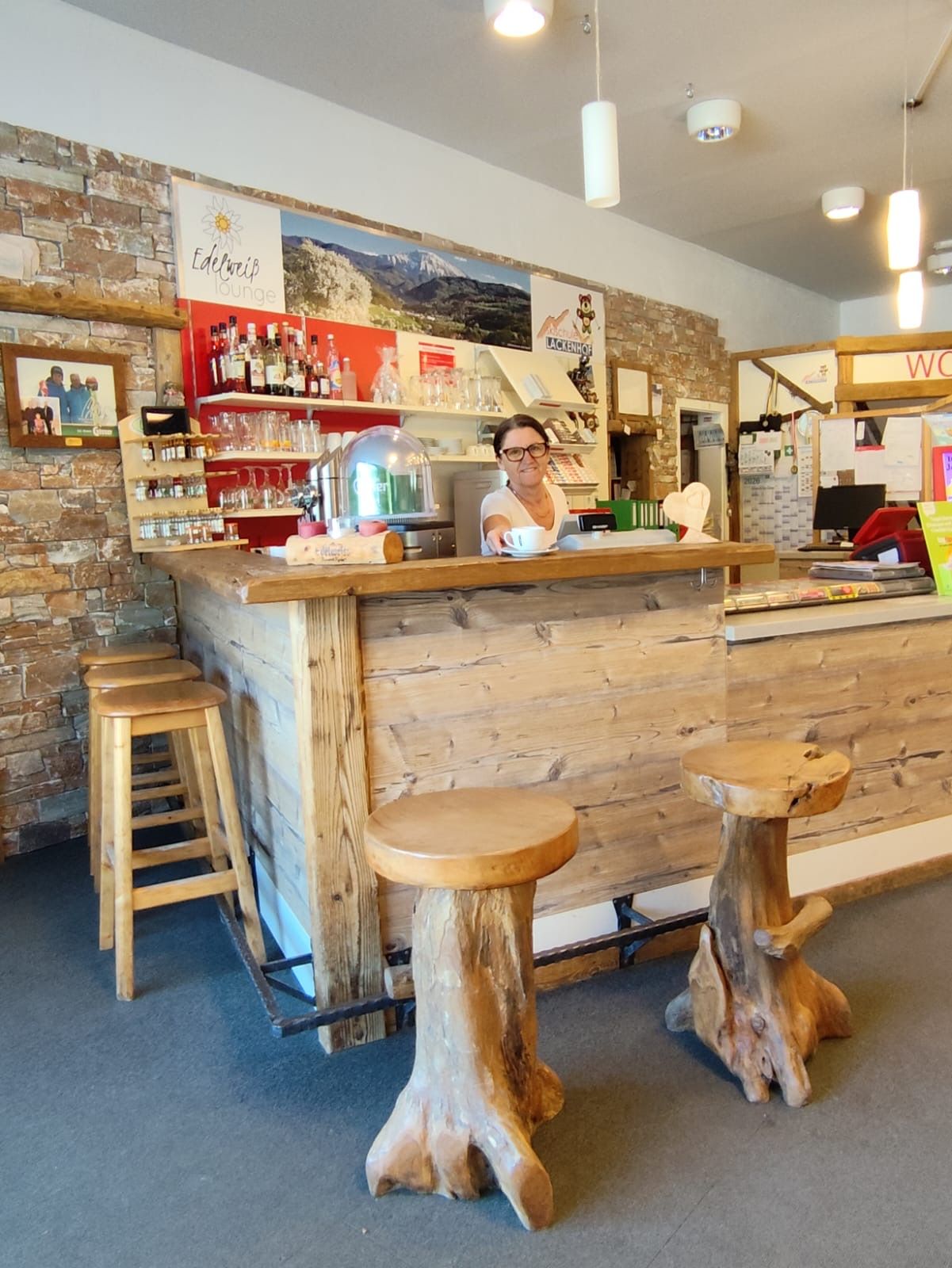 Eine Frau steht hinter einer rustikalen Holztheke in einer gemütlichen Lounge mit Holzhockern und Steinwand.