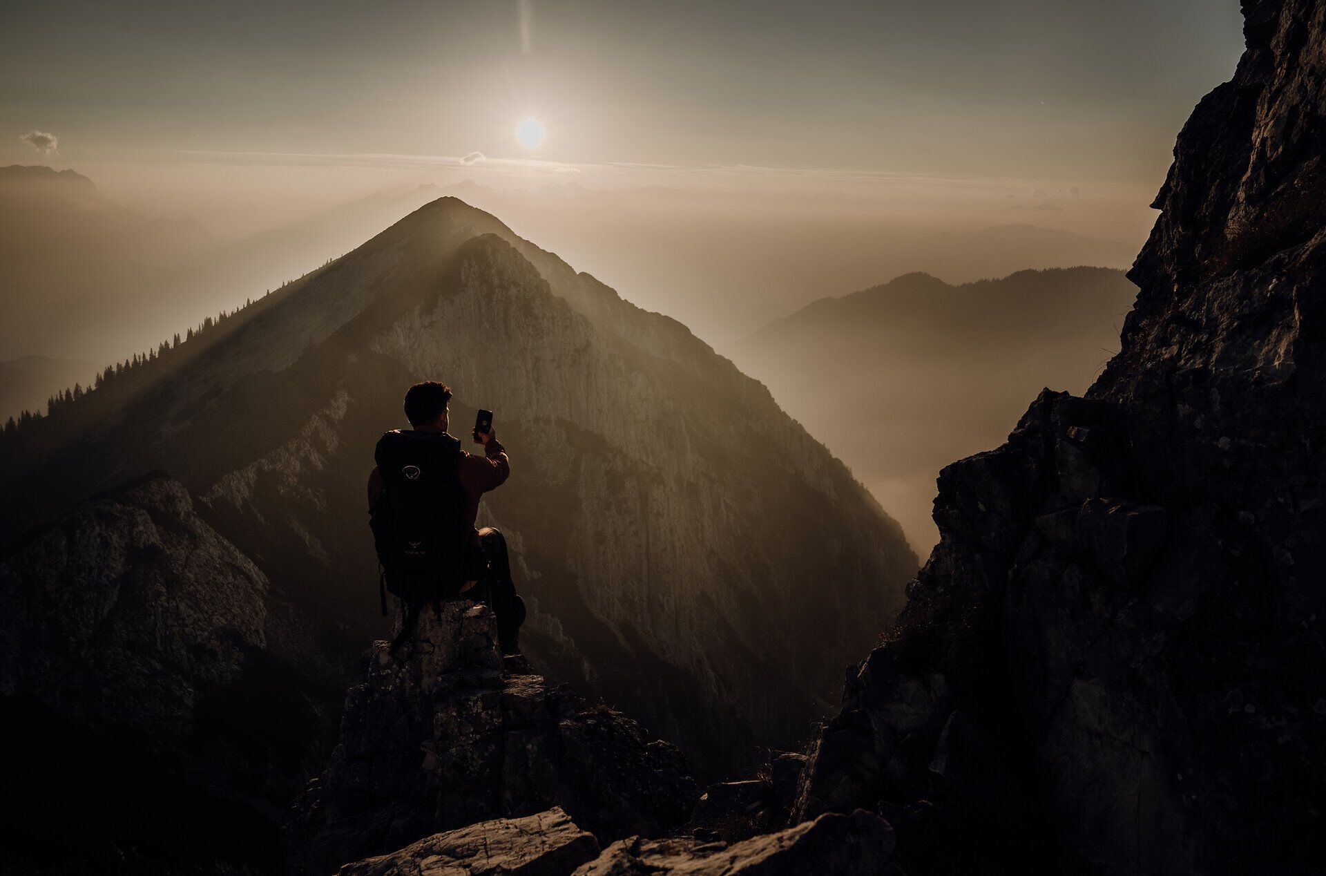 Ein Wanderer sitzt auf einem Felsen und genießt den atemberaubenden Blick auf die sanften Hügel und die untergehende Sonne. Die goldenen Strahlen des Sonnenuntergangs tauchen die Landschaft in ein warmes Licht und schaffen eine friedliche Atmosphäre, die zum Verweilen einlädt.
