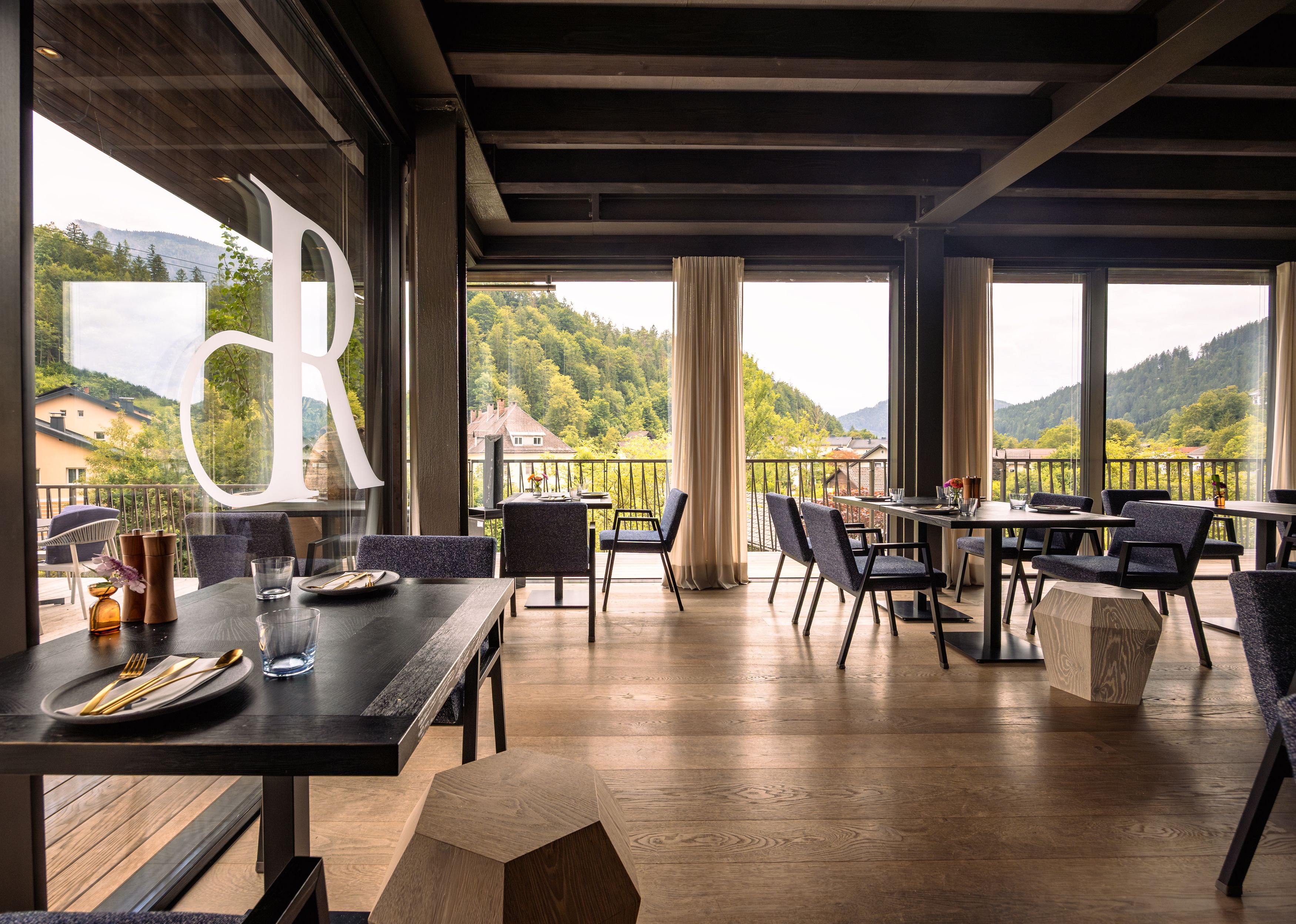 Innenansicht eines modernen Restaurants mit großen Fenstern und Blick auf bewaldete Hügel.