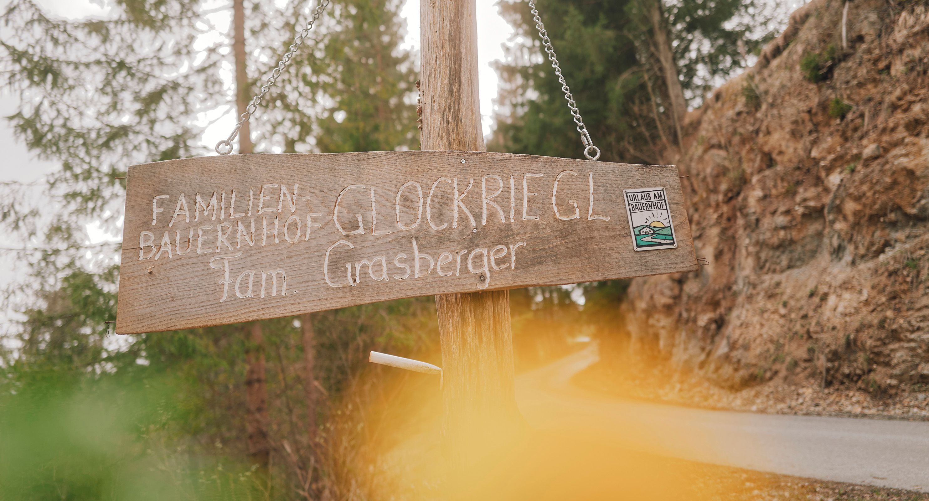 Holzschild mit der Aufschrift 'Familienbauernhof Glockriegl Fam. Grasberger' an einem Waldweg.