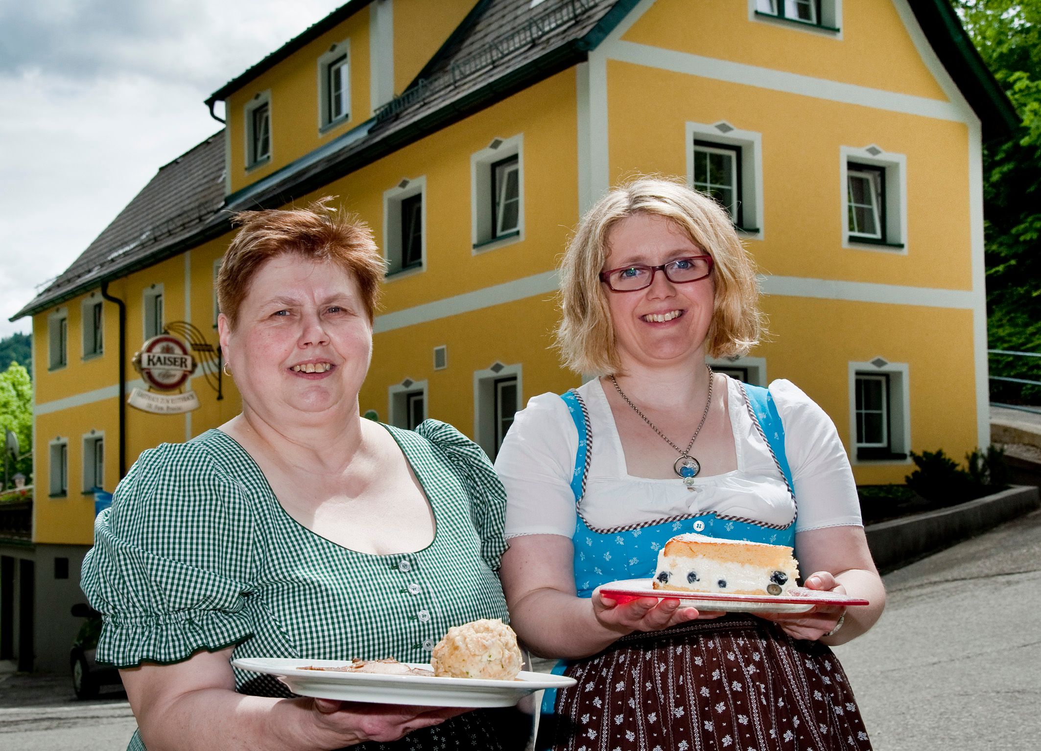 Zwei Frauen in traditioneller Kleidung halten Teller mit Speisen vor einem gelben Gebäude.