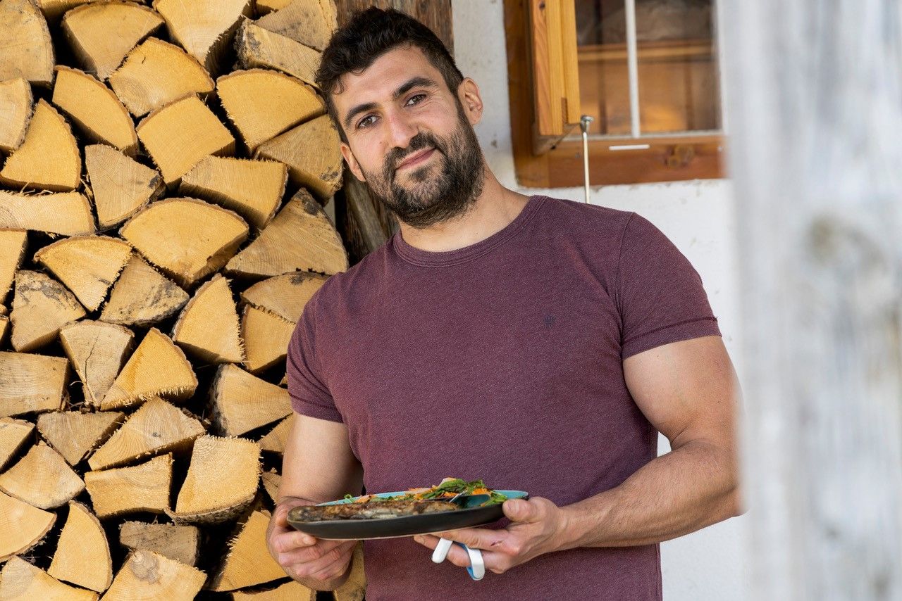 Ein Mann in einem weinroten T-Shirt hält einen Teller mit Essen vor einem Stapel Holzscheite.