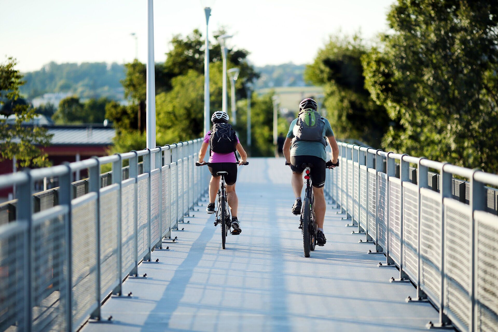 Ein Paar radelt entspannt über eine moderne Brücke, umgeben von üppigem Grün und der sanften Schönheit der Natur. Die warmen Sonnenstrahlen tauchen die Szenerie in ein goldenes Licht und laden dazu ein, die frische Luft und die malerische Landschaft zu genießen.