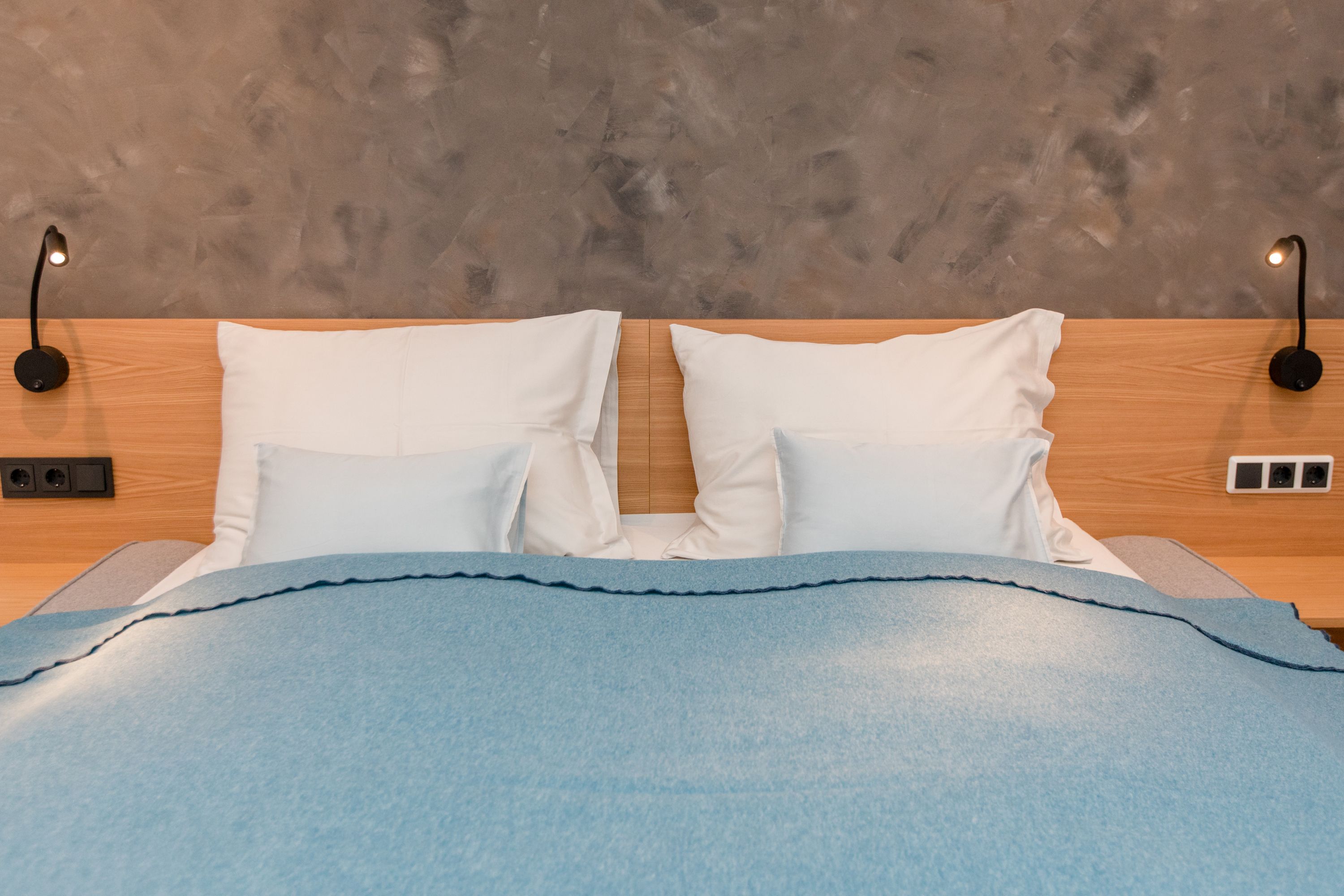 Ein Doppelbett mit blauer Decke und weißen Kissen vor einer grauen Wand.