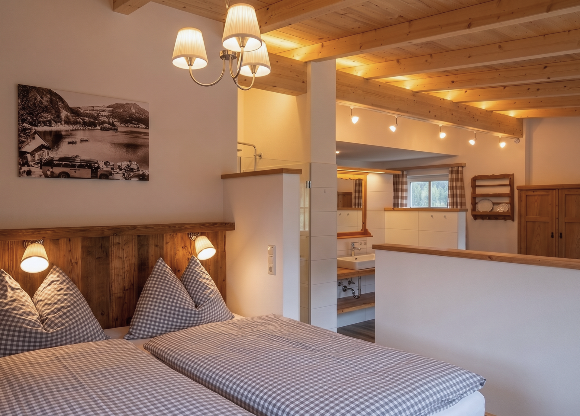 Gemütliches Schlafzimmer mit Holzbalkendecke und Doppelbett, dekoriert mit karierten Kissen und Decken. Im Hintergrund ein Badezimmer mit Waschbecken, hinter der halbhohen Wand befindet sich eine frei stehende Badewanne.